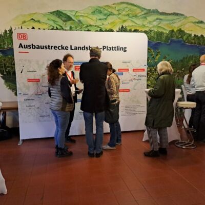 Eine Menschengruppe unterhält sich vor einem Info-Plakat